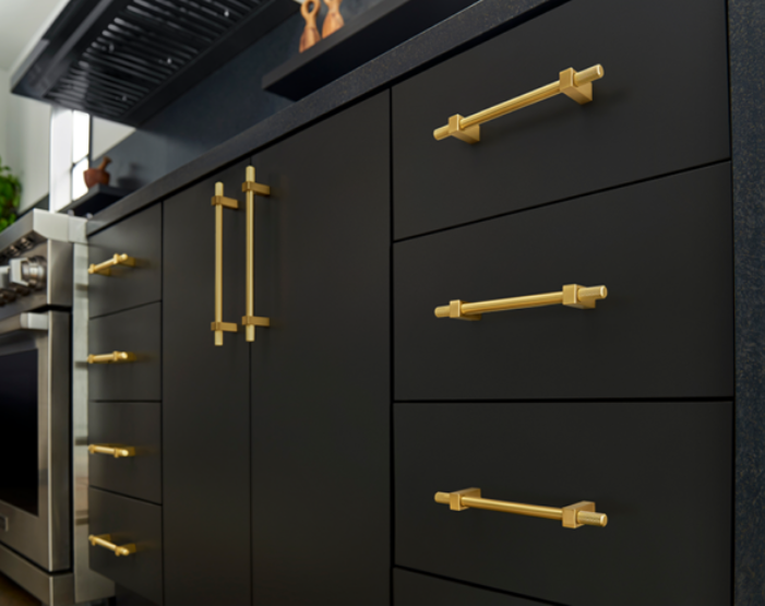 Close-up of dark flat-panel cabinets with polished brass pulls, showcasing a modern and elegant design. A black countertop and subtle background elements complete the sophisticated look.